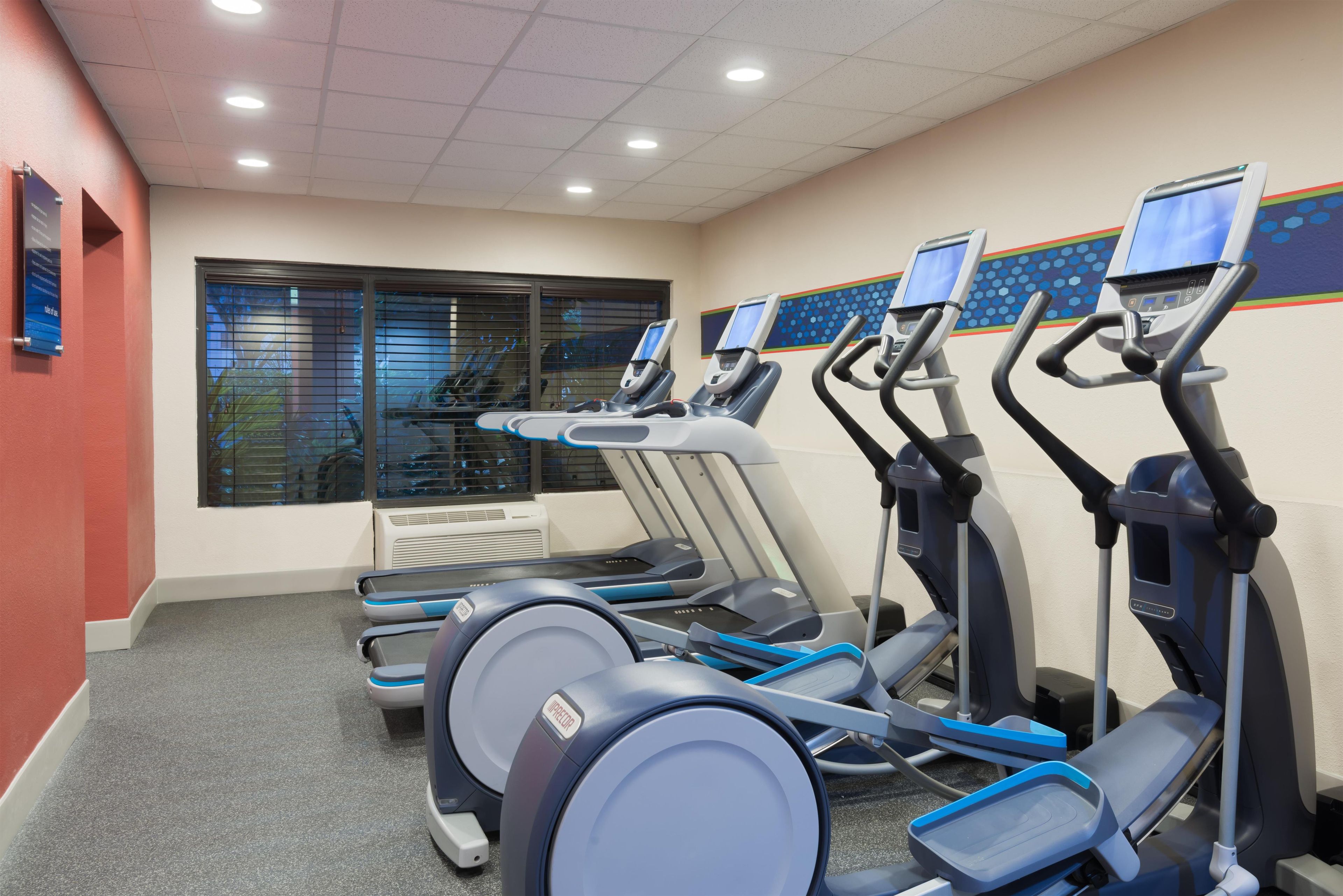Un gimnasio con cintas de correr y elípticas, una ventana con persianas, y una pared roja.