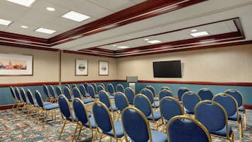 Un salón de conferencias con filas de sillas azules, una alfombra estampada y un techo con iluminación empotrada y detalles de madera.