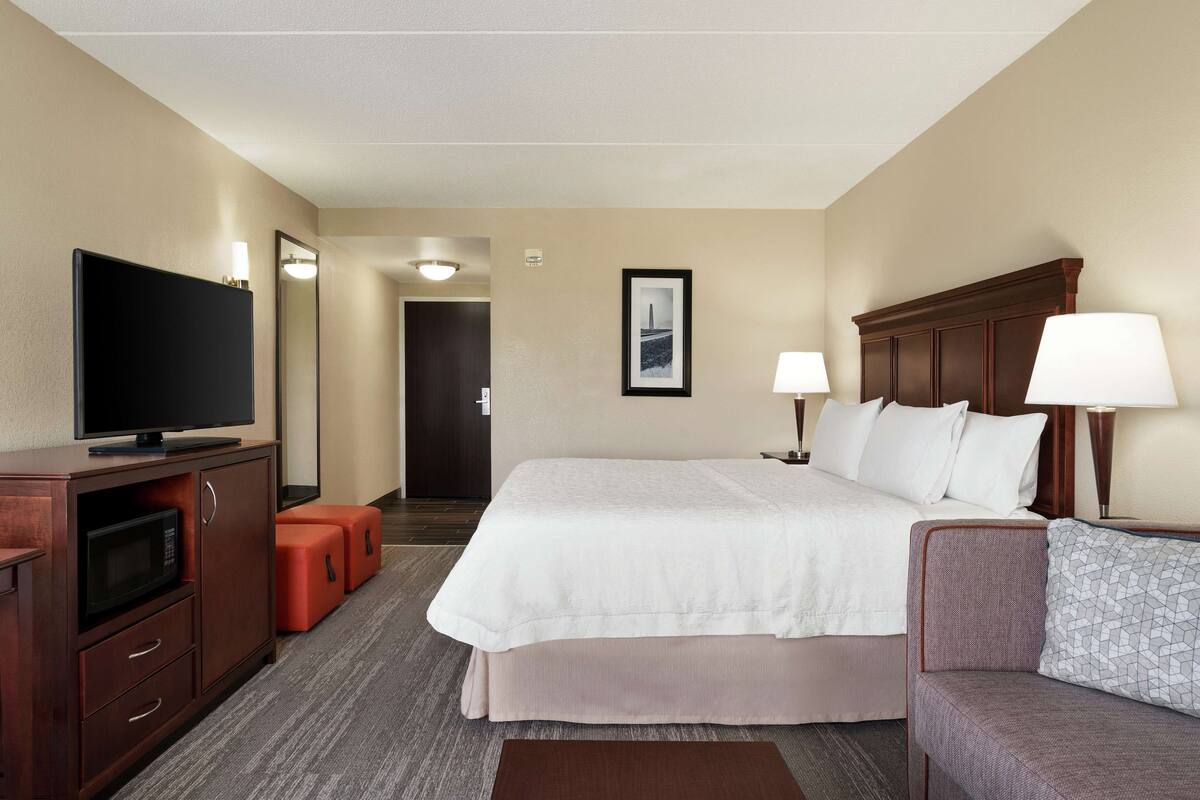 Ein Hotelzimmer mit einem großen Bett, einem Fernseher, einem Schreibtisch mit einer Lampe und einer kleinen Sitzecke.