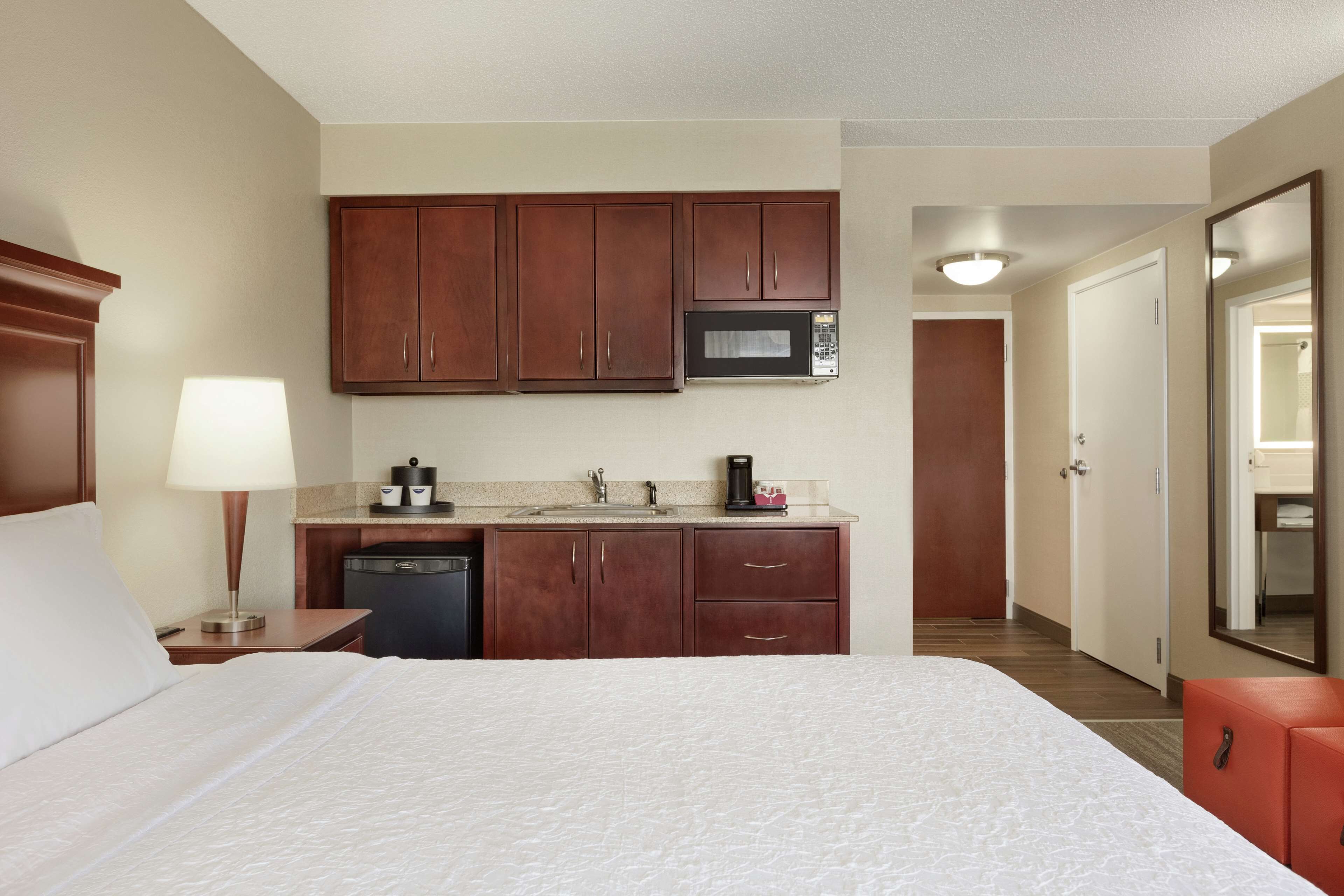A hotel room with a bed, a desk with a lamp, a microwave, a coffee maker, and a small red cabinet.