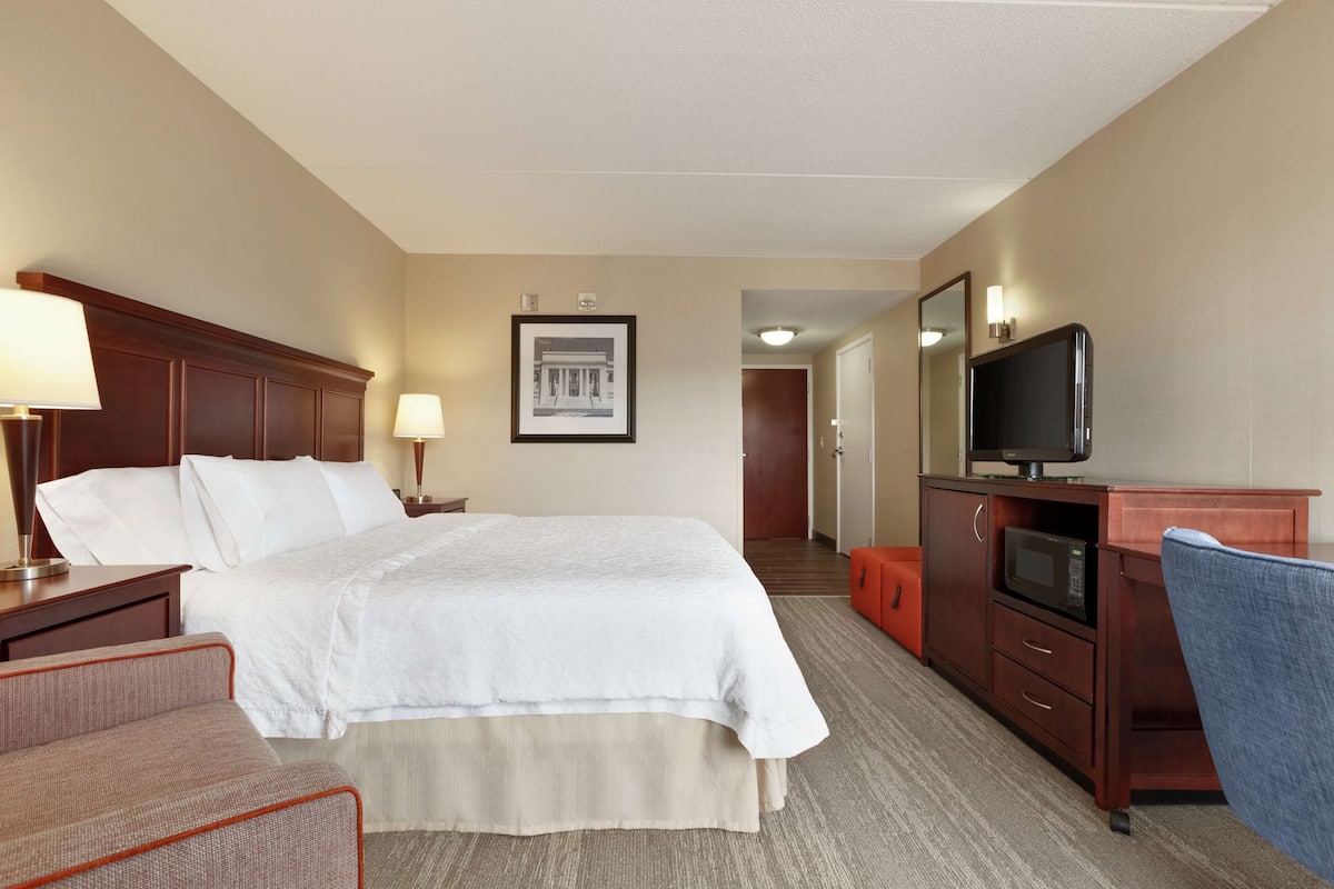 Ein Hotelzimmer mit einem großen Bett, einem Schreibtisch mit Fernseher, einem Sessel und einem gerahmten Bild an der Wand.