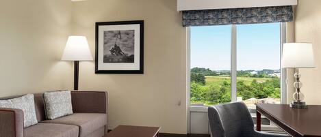 A hotel room with a sofa, armchair, and a table. There is a window with a view of greenery and a framed picture on the wall.
