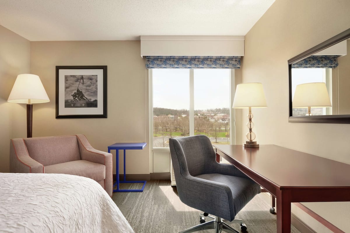 Ein Hotelzimmer mit einem Bett, zwei Sesseln, einem Schreibtisch, einem Fenster mit Blick und einem gerahmten Bild an der Wand.