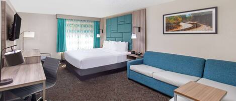 Una habitación de hotel con una cama, un escritorio con silla, un sofá azul, una mesita y un cuadro en la pared.