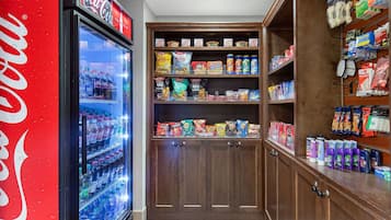 A vending machine stocked with Coca-Cola and various snacks, surrounded by shelves with packaged goods and toiletries.