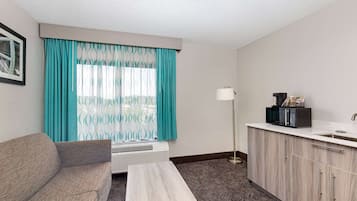 Una habitación de hotel compacta con sofá, una mesita, kitchenette con microondas y ventana con cortinas estampadas.