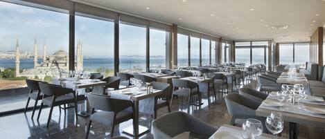 A restaurant with a long dining area, large windows, and a view of a cityscape and sea.