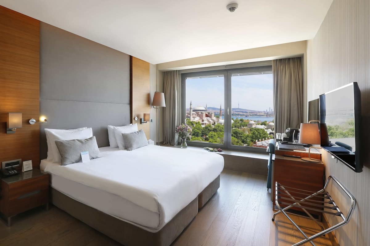 A hotel room with a large bed, bedside tables, a desk with a chair, a TV, and a view of a cityscape.