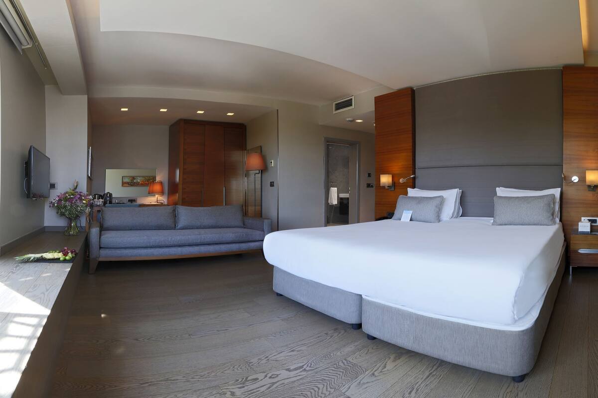 A modern hotel room with a large bed, a sofa, and a view of greenery through large windows.