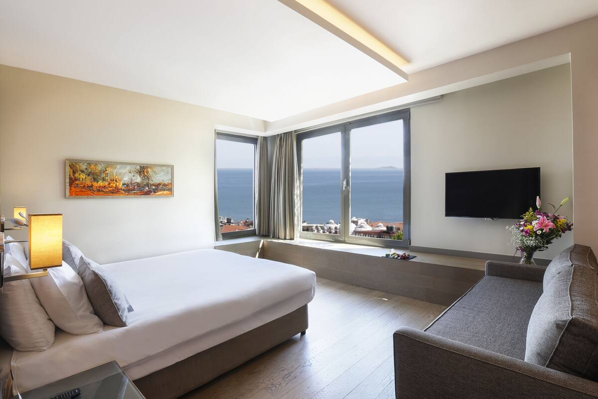 A modern hotel room with a bed, a sofa, a TV, and a large window offering a view of the sea.