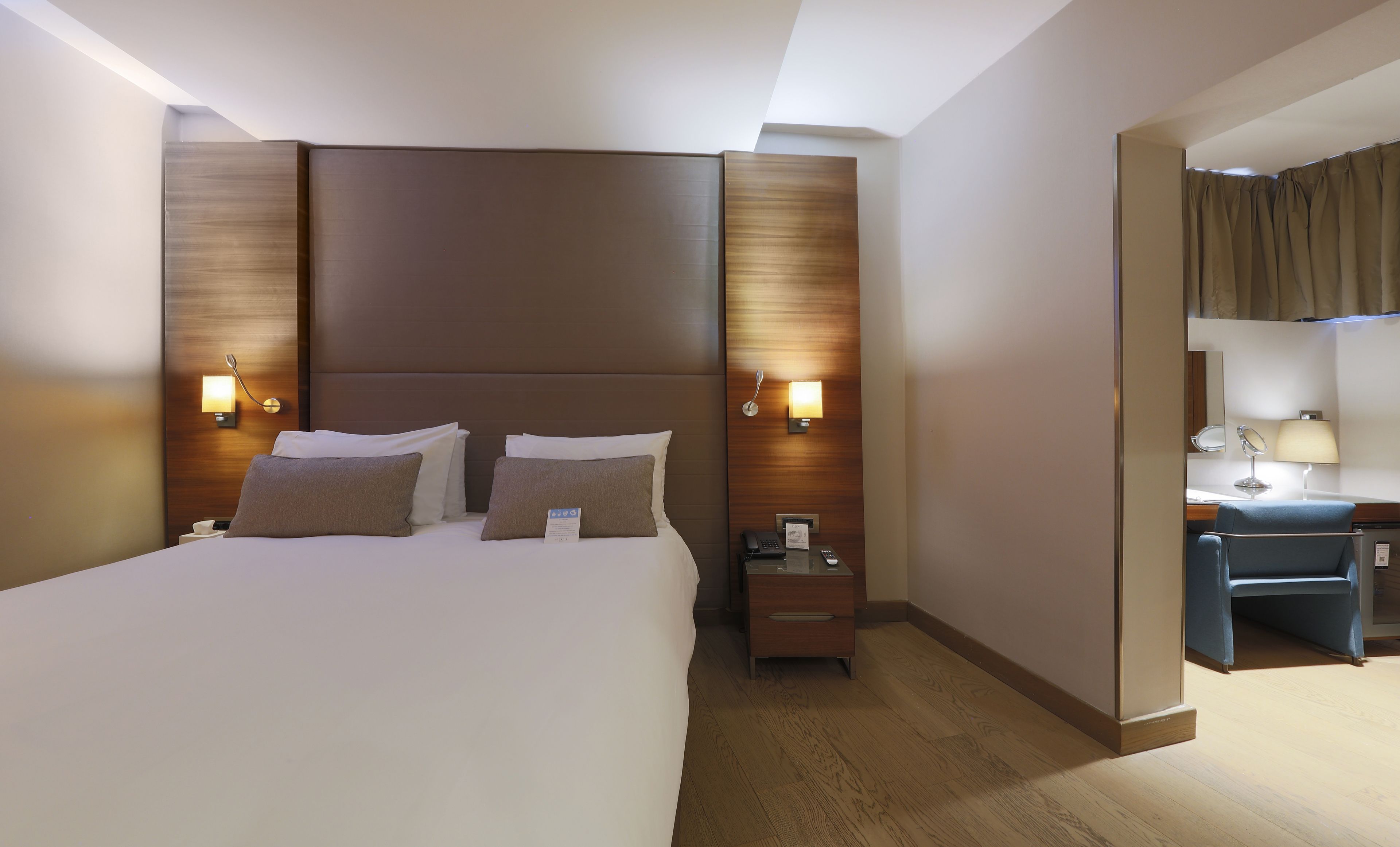 A hotel room with a large bed, bedside lamps, a nightstand, a desk with a chair, and a mirror.
