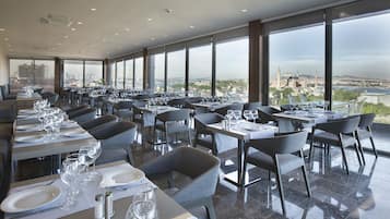 Restaurante com várias mesas preparadas para refeições, grandes janelas com vista para a cidade e design de interiores moderno.