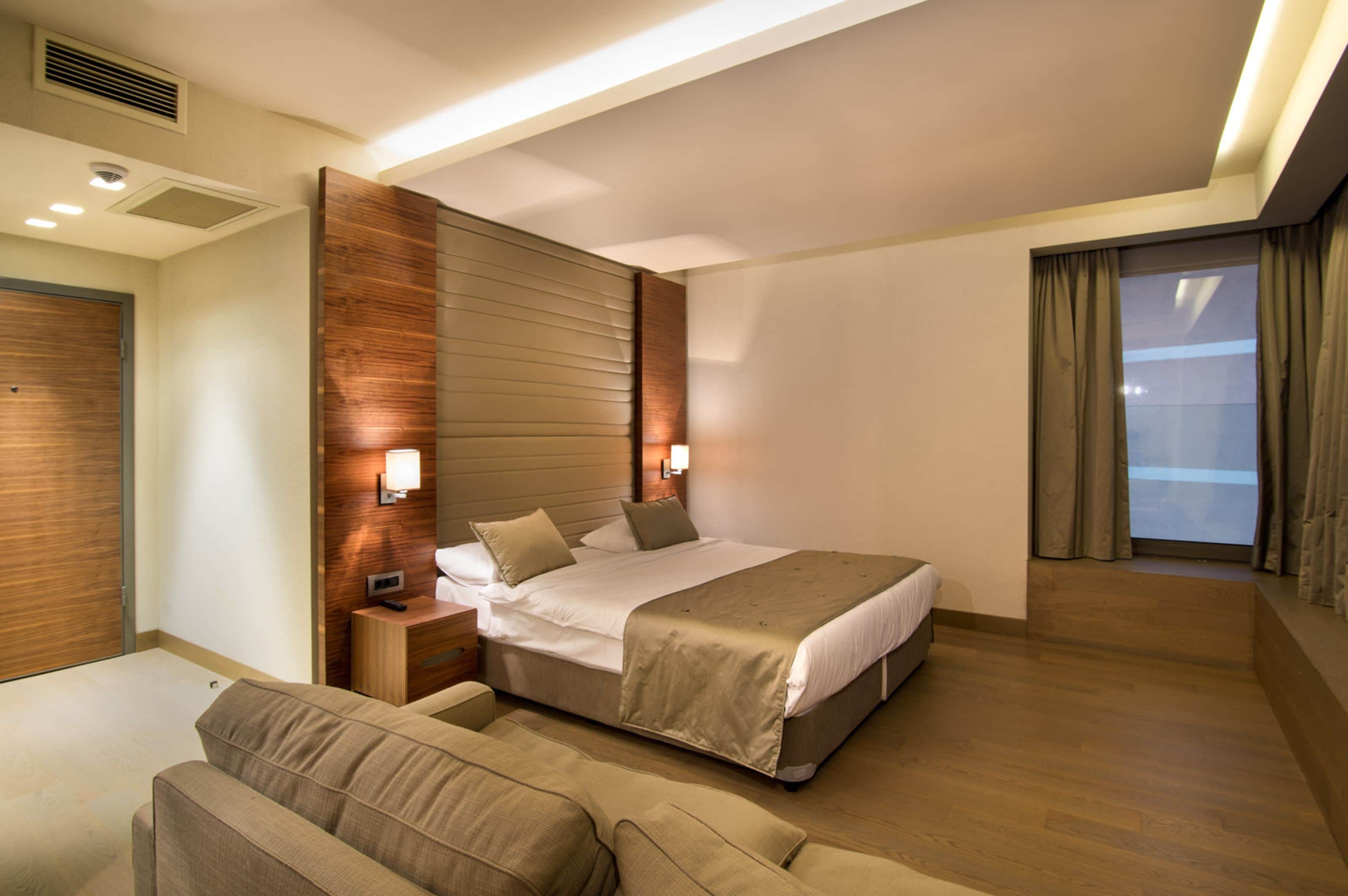 A modern hotel room with a large bed, a bedside table, a nightstand, a sofa, and a large window with curtains.