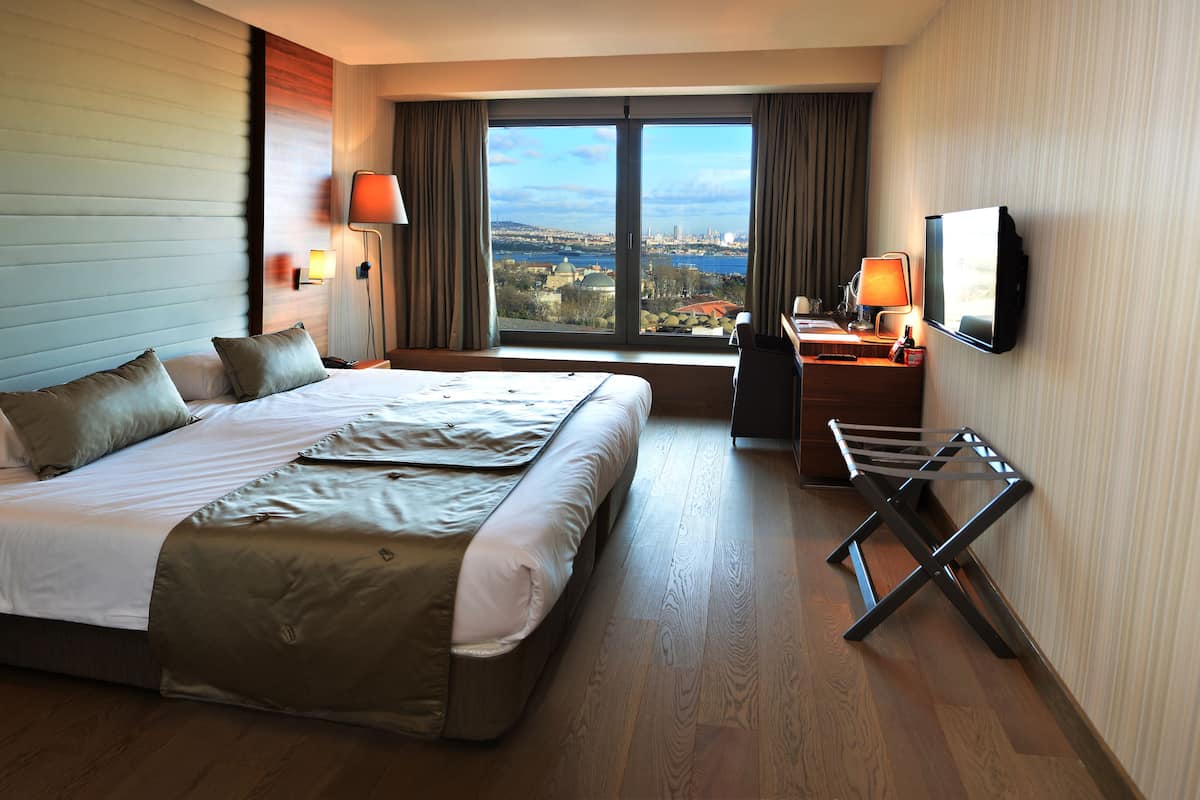 A hotel room with a large bed, a desk with a lamp, a TV, and a view of a cityscape through a large window.