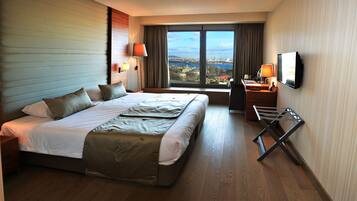 A hotel room with a large bed, a desk with a lamp, a TV, and a view of a cityscape through a large window.