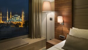 A hotel room with a large window overlooking a cityscape at night, a bedside lamp, a wooden headboard, and a bed with pillows. - Hotel Arcadia Blue Istanbul (Istanbul)