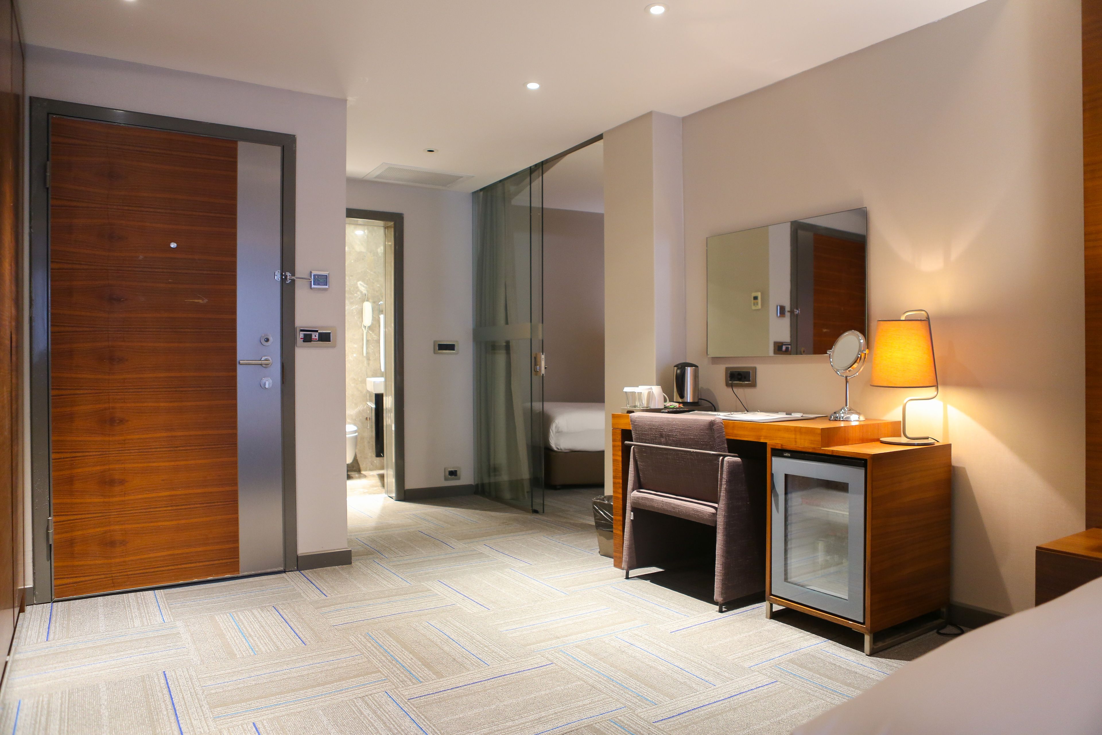 A hotel room with a wooden door, a desk with a lamp, a mirror, and a bed.