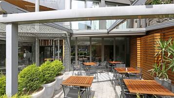 An outdoor cafe area with multiple tables and chairs, surrounded by potted plants and a modern architectural design.