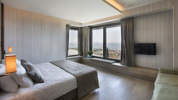 Ein Hotelzimmer mit einem großen Bett, einem an der Wand montierten Fernseher und Blick auf Stadt und Meer durch die Fenster.