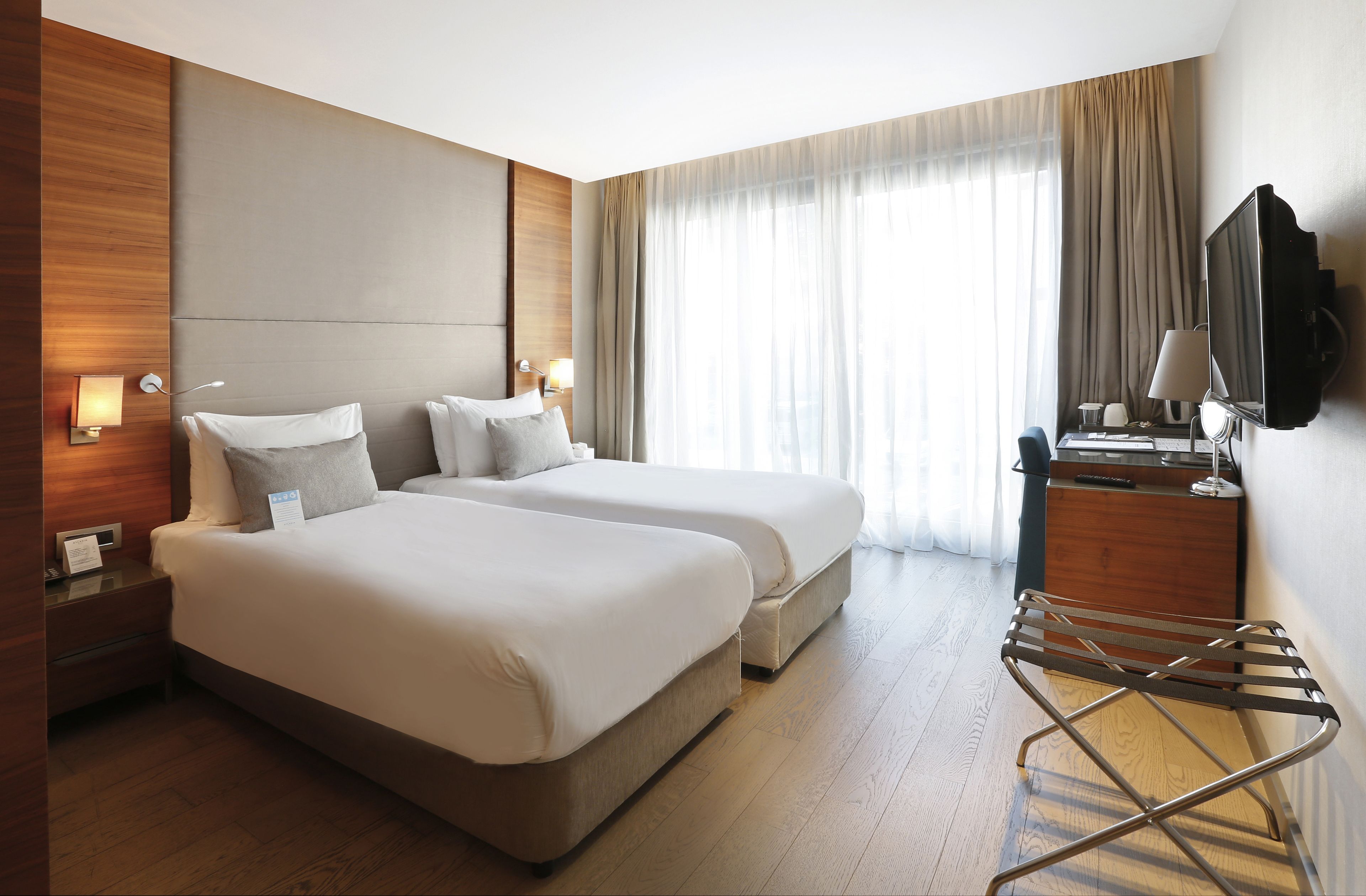 A hotel room with two beds, a desk with a computer, a TV, and a window with curtains.