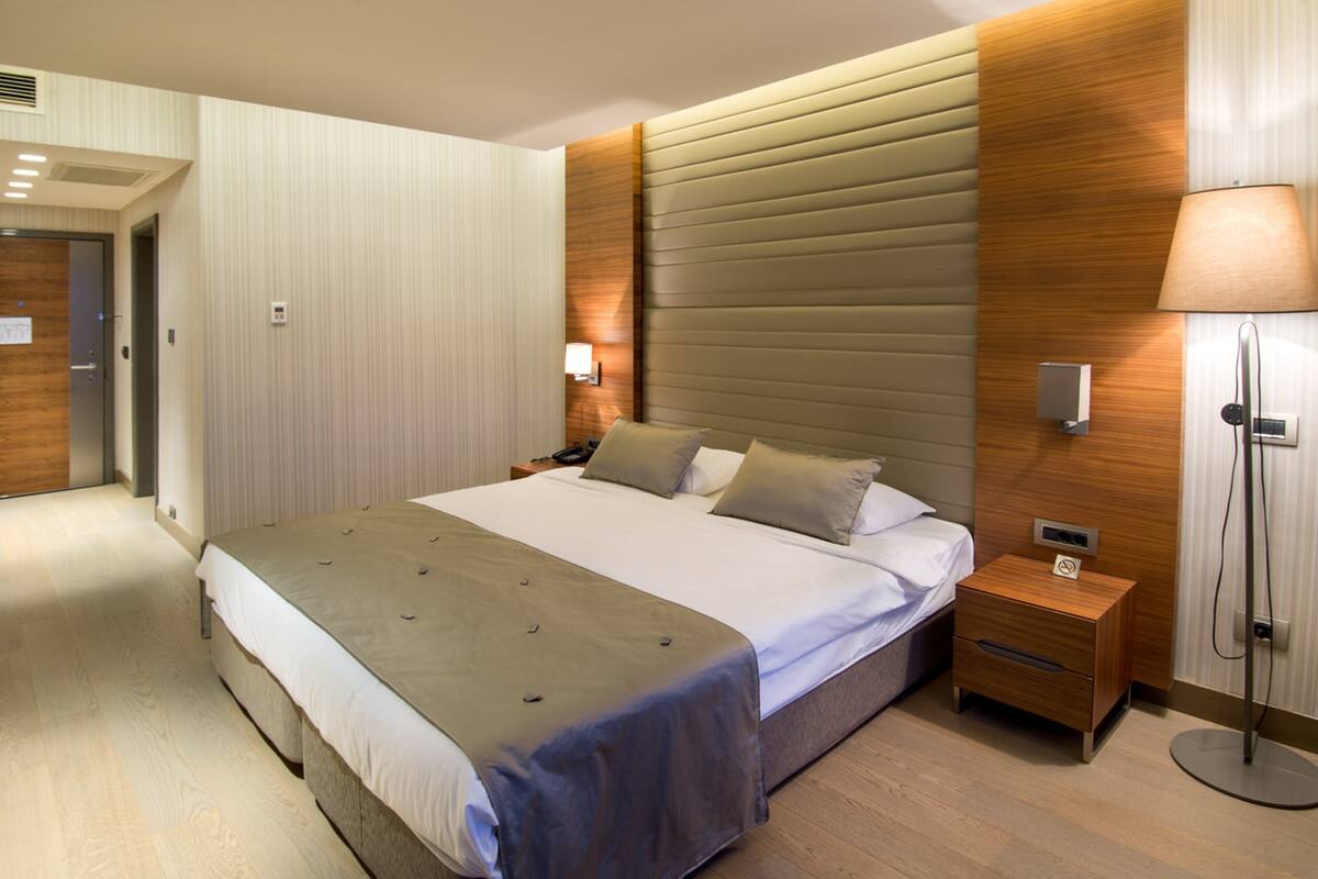 A modern hotel room with a large bed, a bedside table, a nightstand, a lamp, and a wall-mounted light fixture.