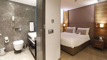 Una habitación de hotel moderna con un baño de mármol, un dormitorio independiente con una cama grande y un cabecero de madera.