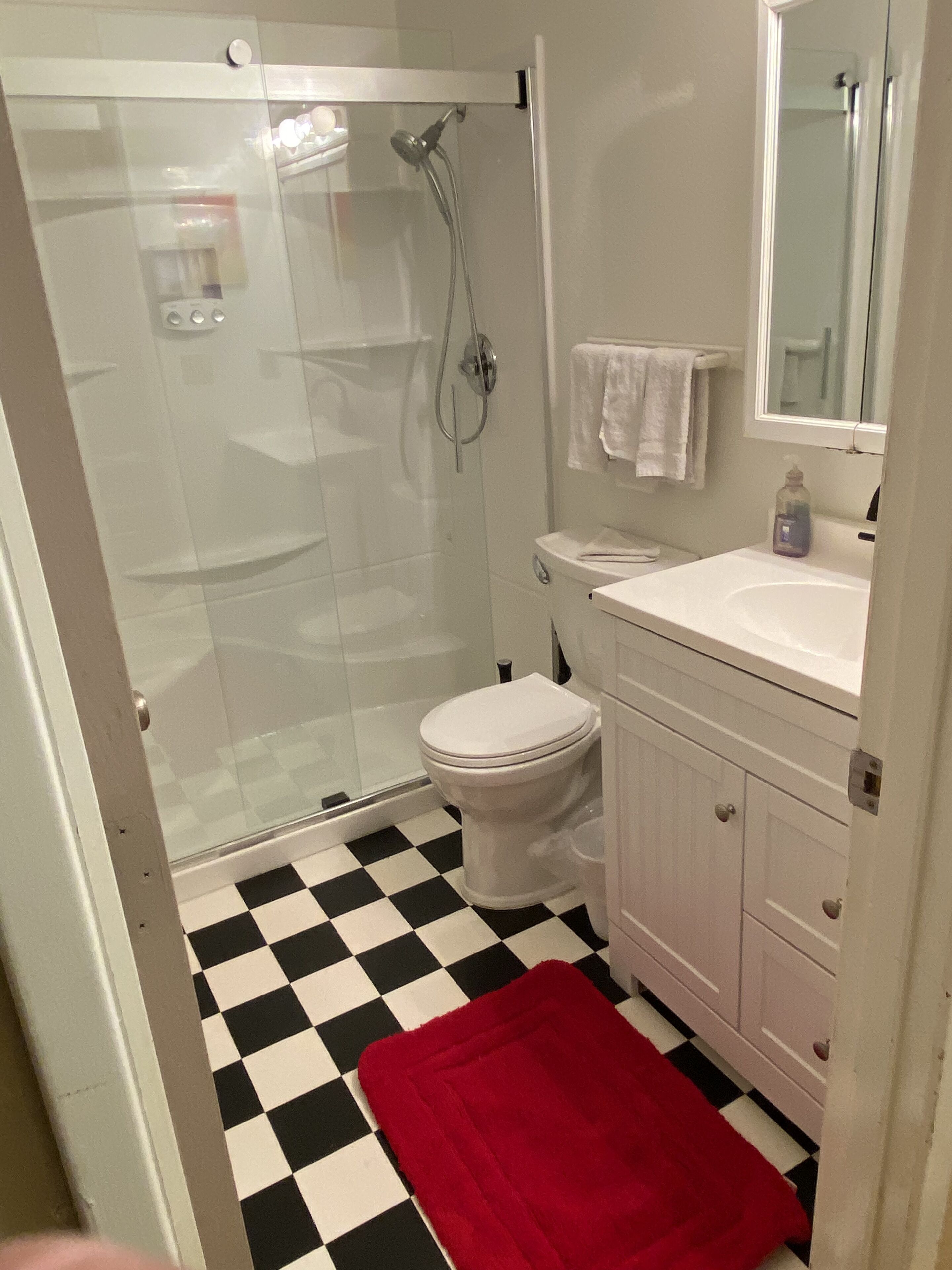 A bathroom with a glass-enclosed shower, a white toilet, a sink with a mirror above it, and a red bath mat on the floor.