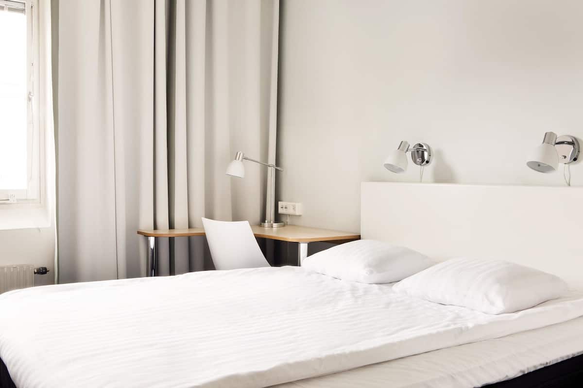 A neatly made bed with white linens, a desk with a lamp, and a chair in a modern hotel room.