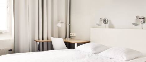Una cama bien hecha con sábanas blancas, un escritorio con lámpara y una silla en una habitación de hotel moderna.