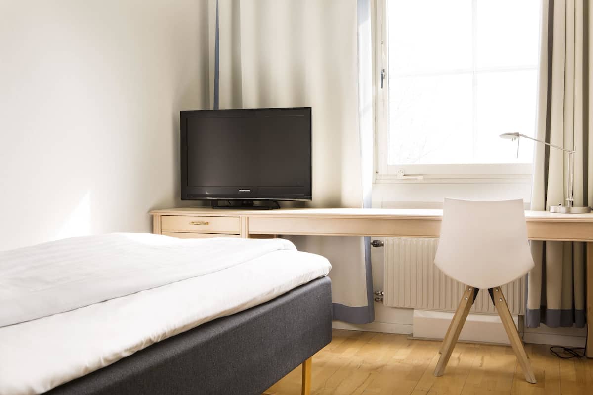 A bedroom with a bed, a desk with a television, a chair, and a window with curtains.