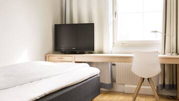 Un dormitorio con cama, escritorio con televisor, silla y ventana con cortinas.