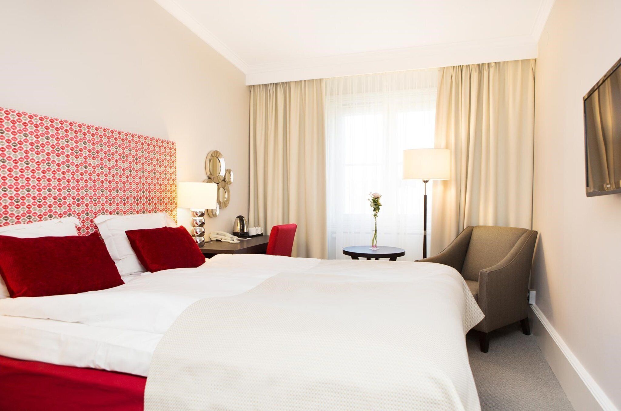A hotel room with a large bed, a desk with a lamp, a chair, and a small table with a vase of flowers.