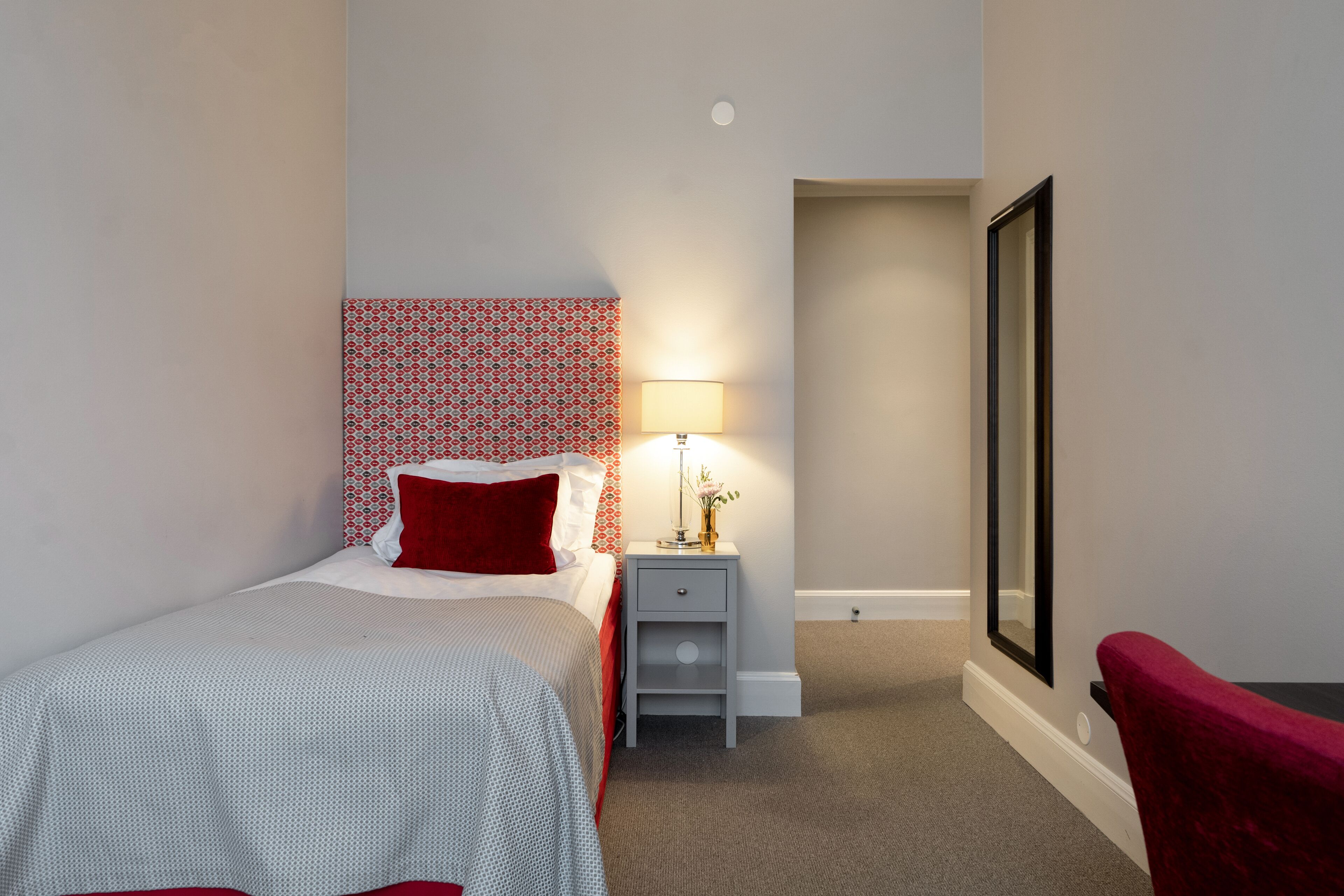 Habitación de hotel con una cama, una mesita de noche con lámpara, un espejo y una silla roja.