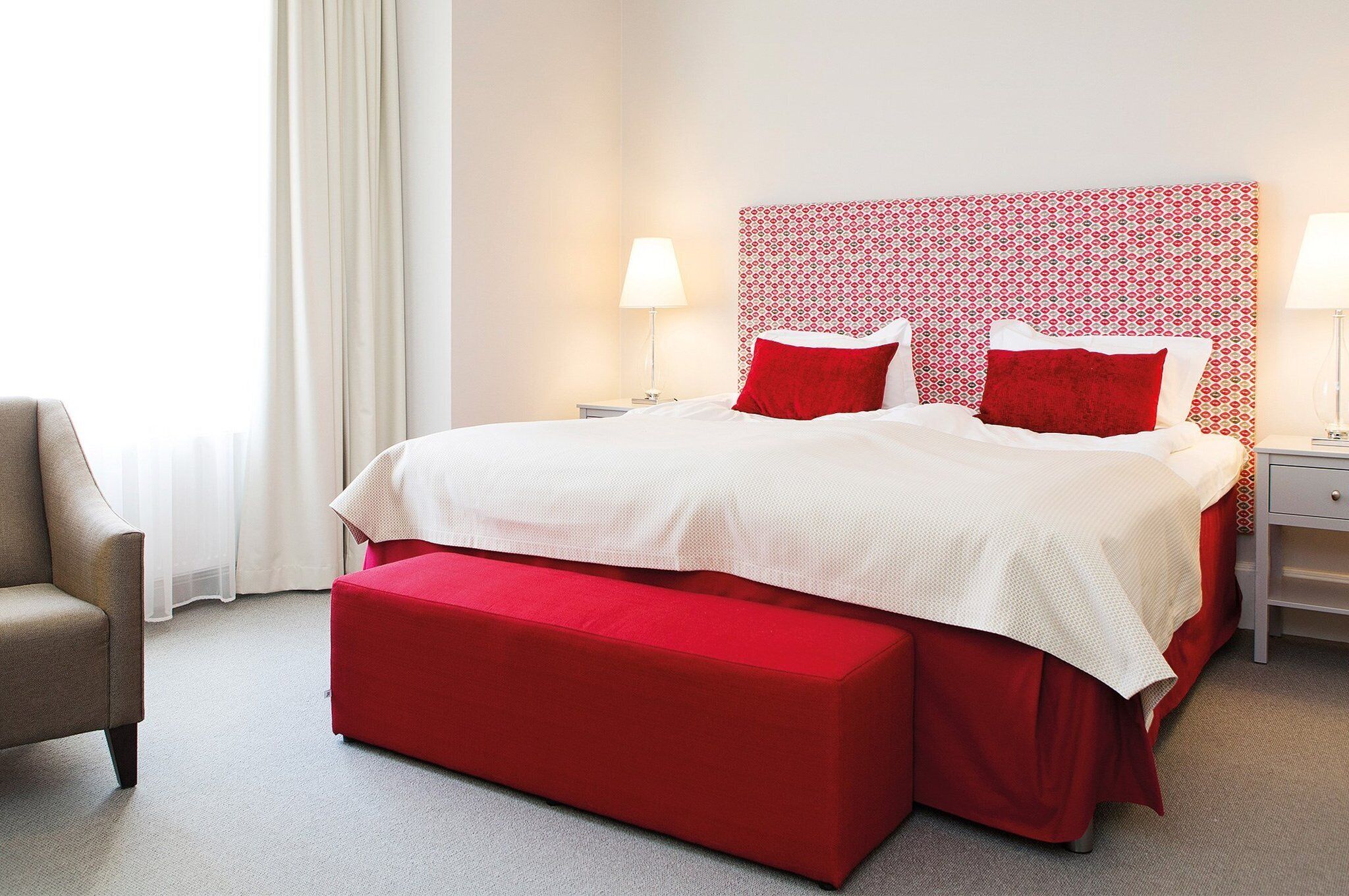 Ein Hotelzimmer mit einem großen Bett, roter Bettwäsche, einer roten Bank, einem gemusterten Kopfteil und zwei Nachttischlampen.
