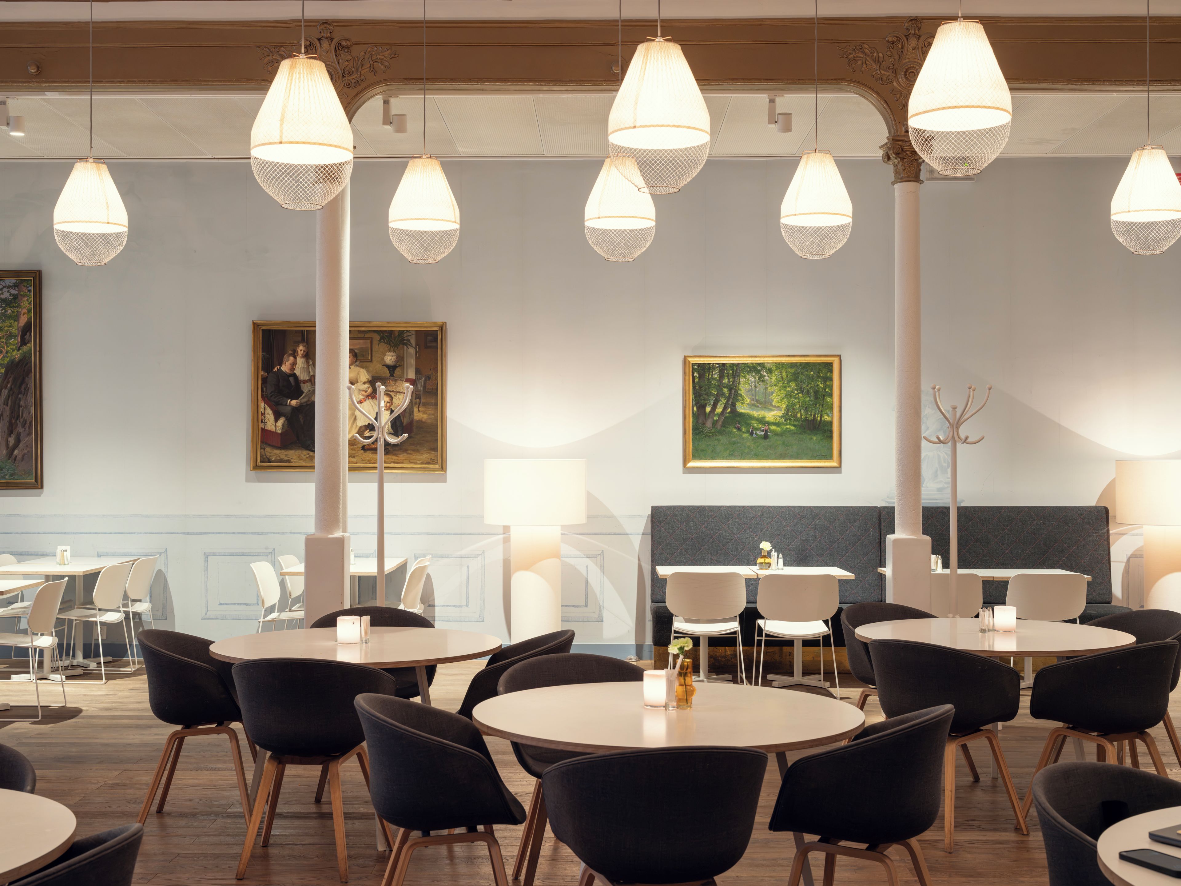 A hotel dining area with round tables, black chairs, hanging pendant lights, and two paintings on the walls.