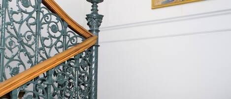 Una escalera con barandillas metálicas ornamentales y un pasamanos de madera, que conduce a un rellano con una araña y un cuadro en la pared.