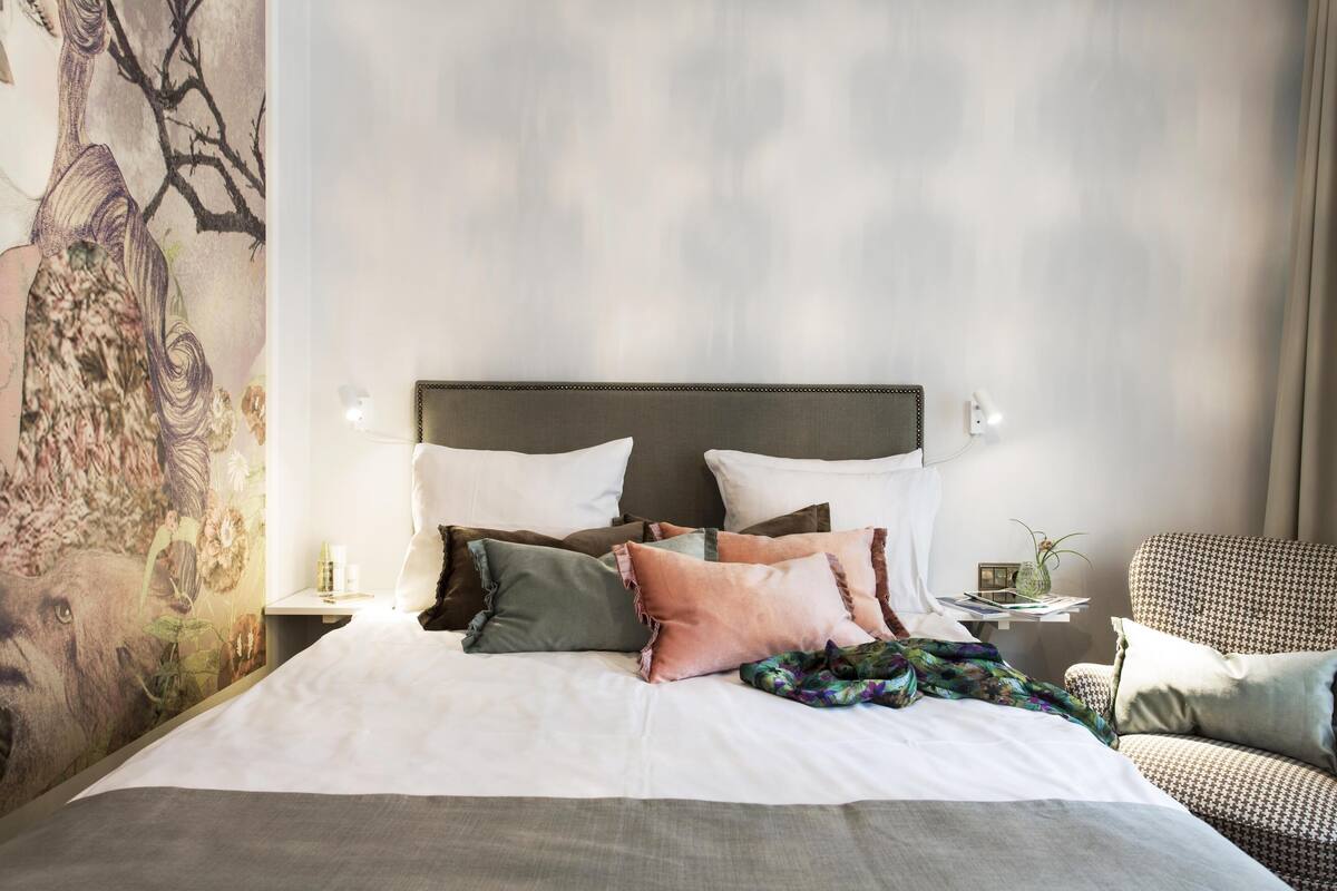 Un letto rifatto con molti cuscini e una coperta, un murales decorativo sulla parete, un comodino con una lampada e una poltrona con motivo.