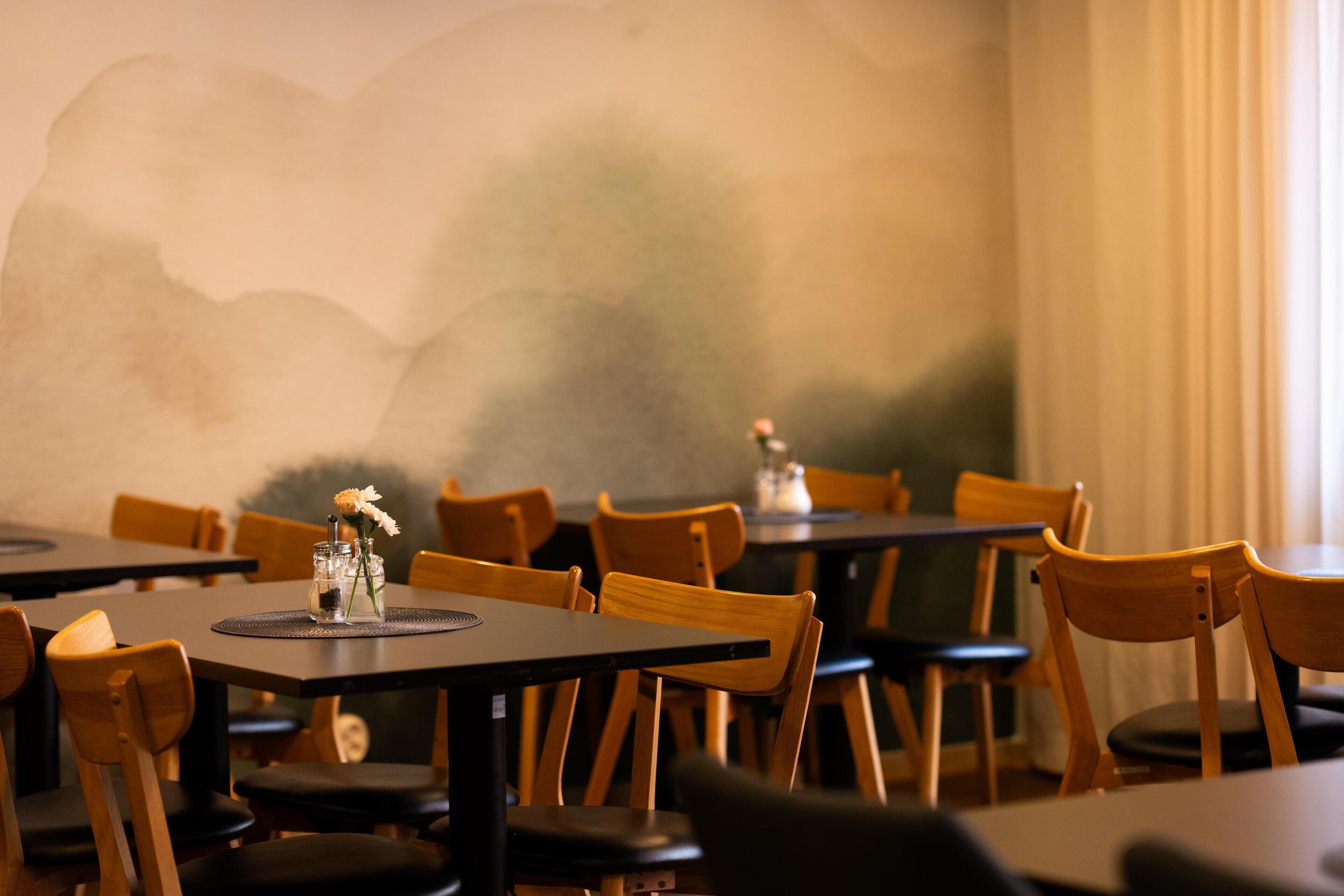 Ein Restaurantinnenraum mit Holztischen und -stühlen, einer Wandmalerei und einer Blumenvase auf den Tischen.