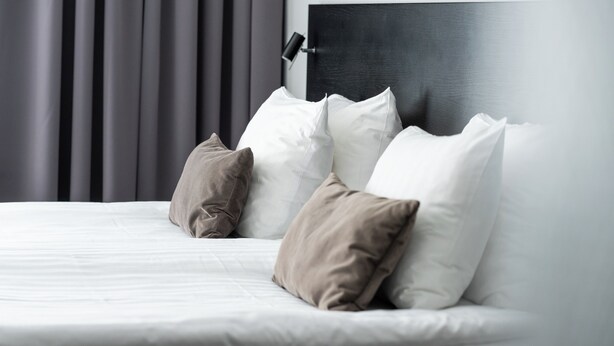 Un lit bien fait, agrémenté d’oreillers blancs et beiges, avec une tête de lit sombre, et une chambre au design moderne et minimaliste.