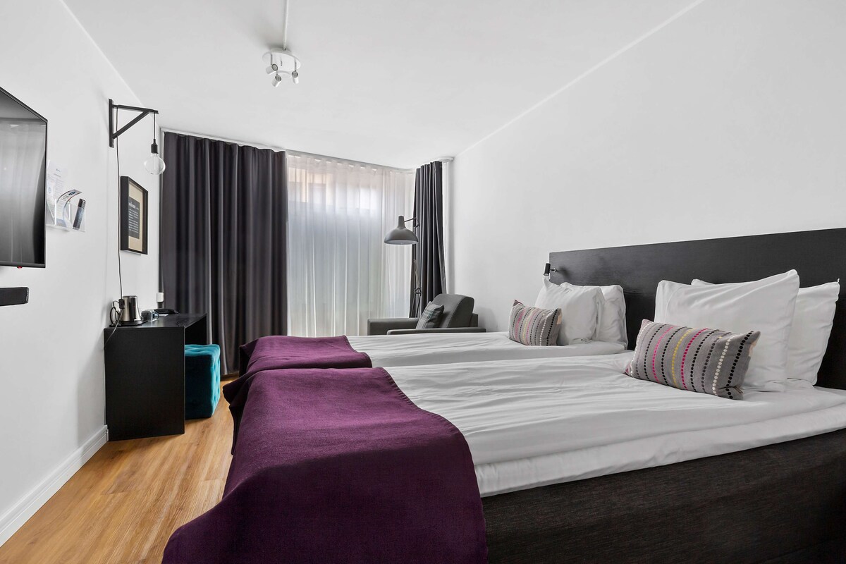 Ein Hotelzimmer mit zwei Betten, einem Schreibtisch, einem Stuhl und einem großen Fenster mit Vorhängen.
