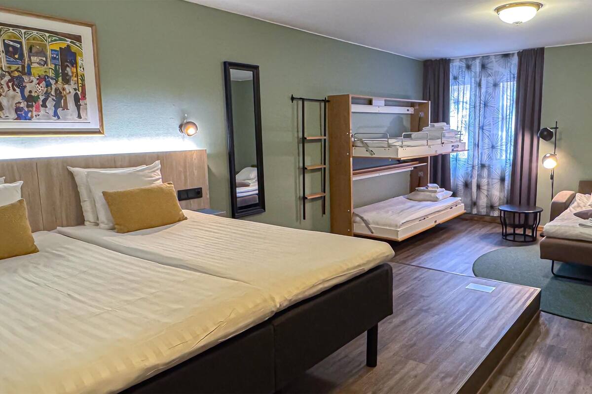Camera d'albergo con due letti, un divano, un letto a castello, uno specchio e un quadro appeso al muro.