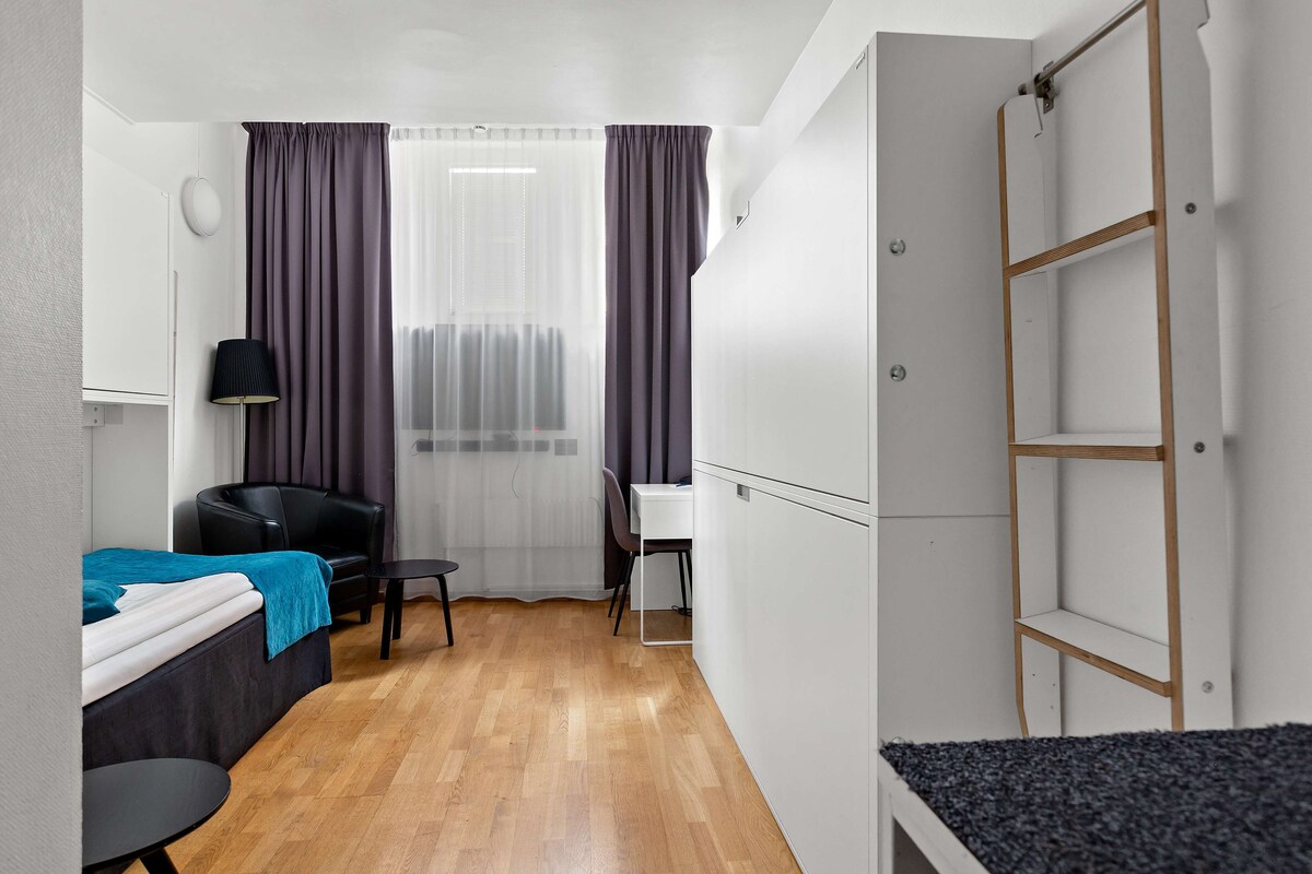 Ein kleines Hotelzimmer mit Bett, Schreibtisch, Stuhl, Kleiderschrank und einem fenster mit Vorhängen.