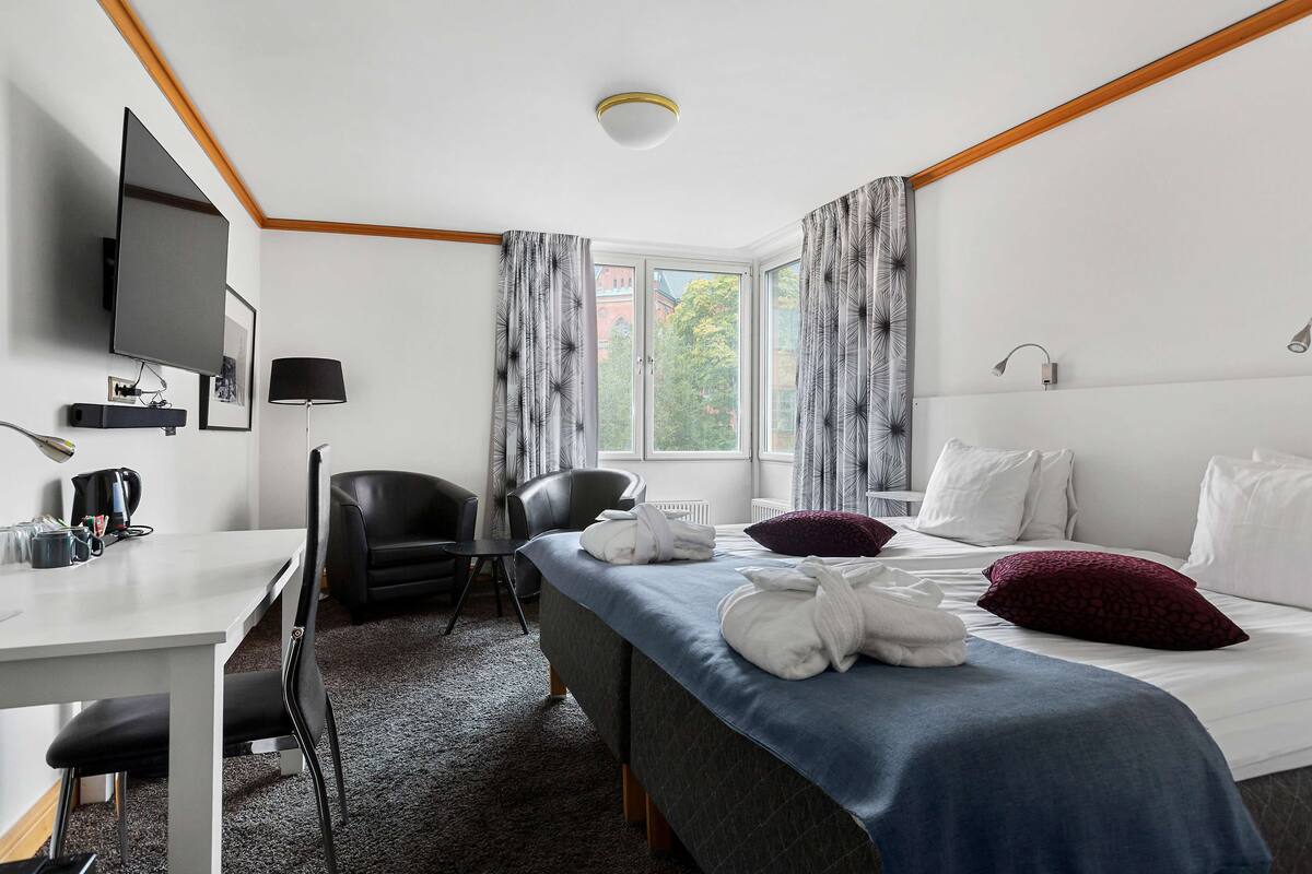 Una camera d'albergo con un letto, una scrivania, una sedia, una televisione e una finestra con tende.
