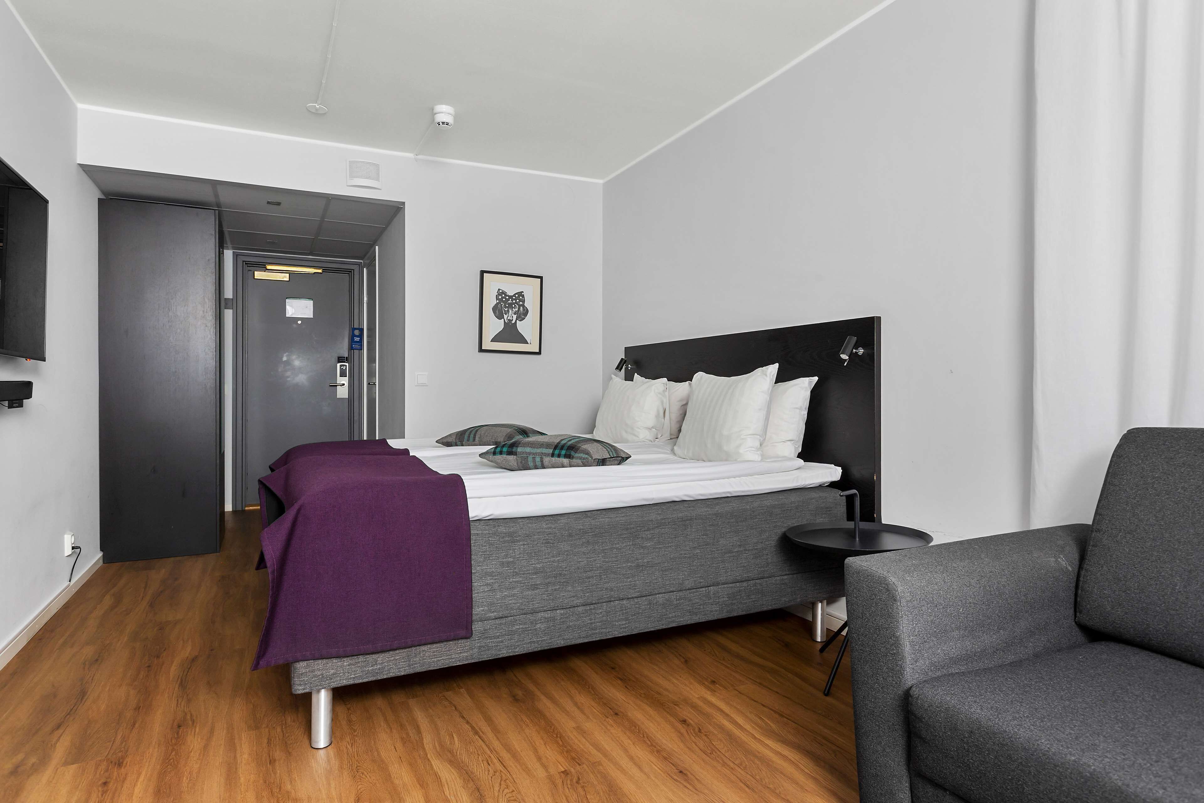 Quarto de hotel moderno com uma cama, uma mesinha, uma cadeira e uma TV fixada na parede.