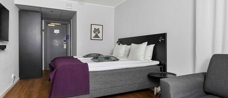 Quarto de hotel moderno com uma cama, uma mesinha, uma cadeira e uma TV fixada na parede.