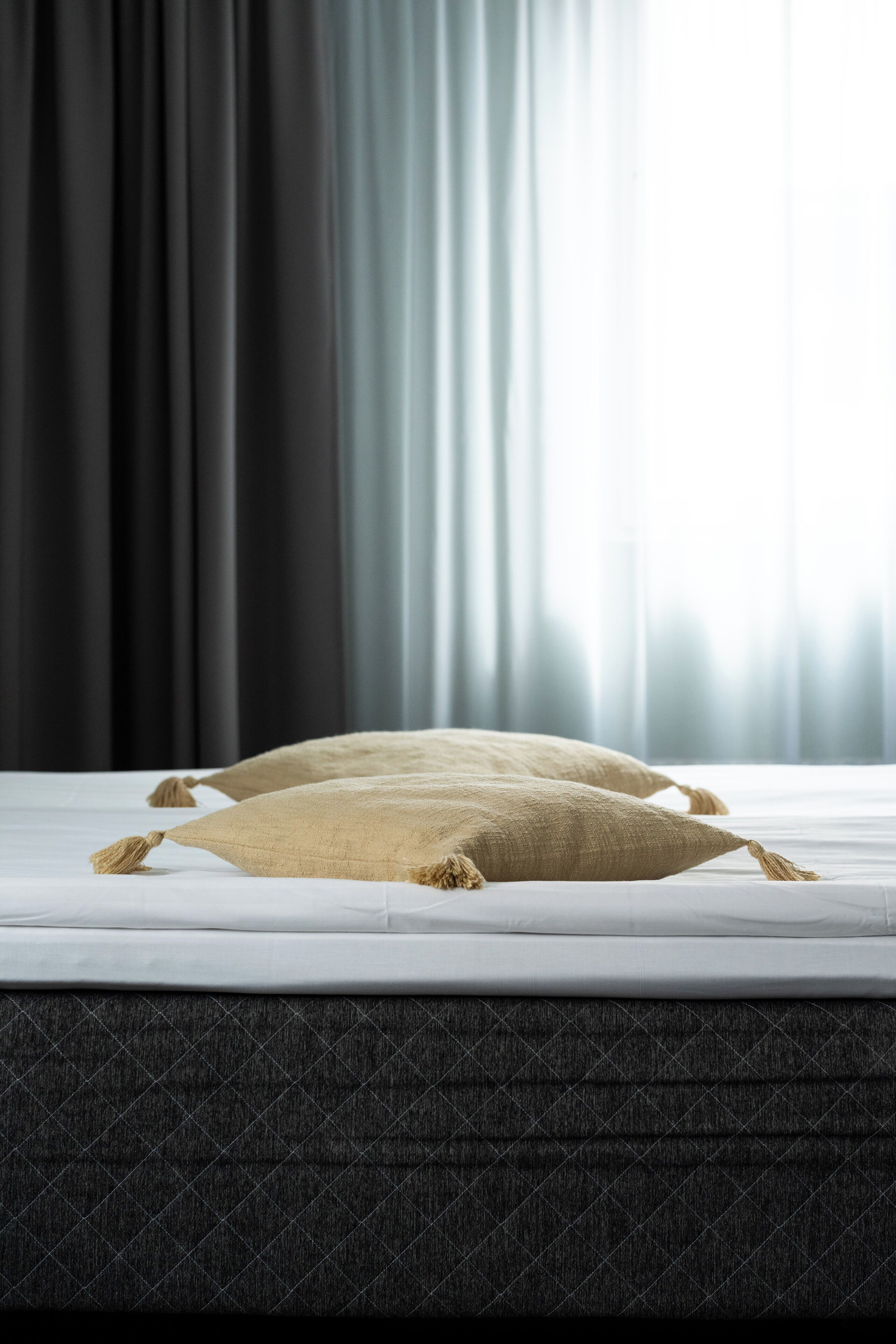 A bed with a plain white mattress, a dark quilted bedspread, and two beige pillows with tassels.