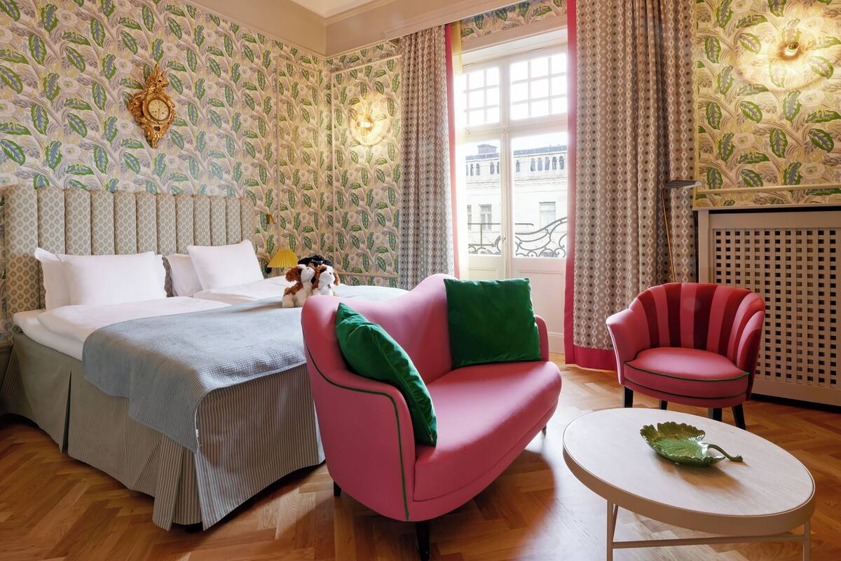 A hotel room with a bed, a pink armchair, a red armchair, and a small white table with a decorative leaf.