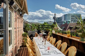 An outdoor dining area with wicker chairs and a long table set for a meal, overlooking a cityscape with buildings and a statue. - Hotel Kung Carl, WorldHotels Crafted (Stockholm)
