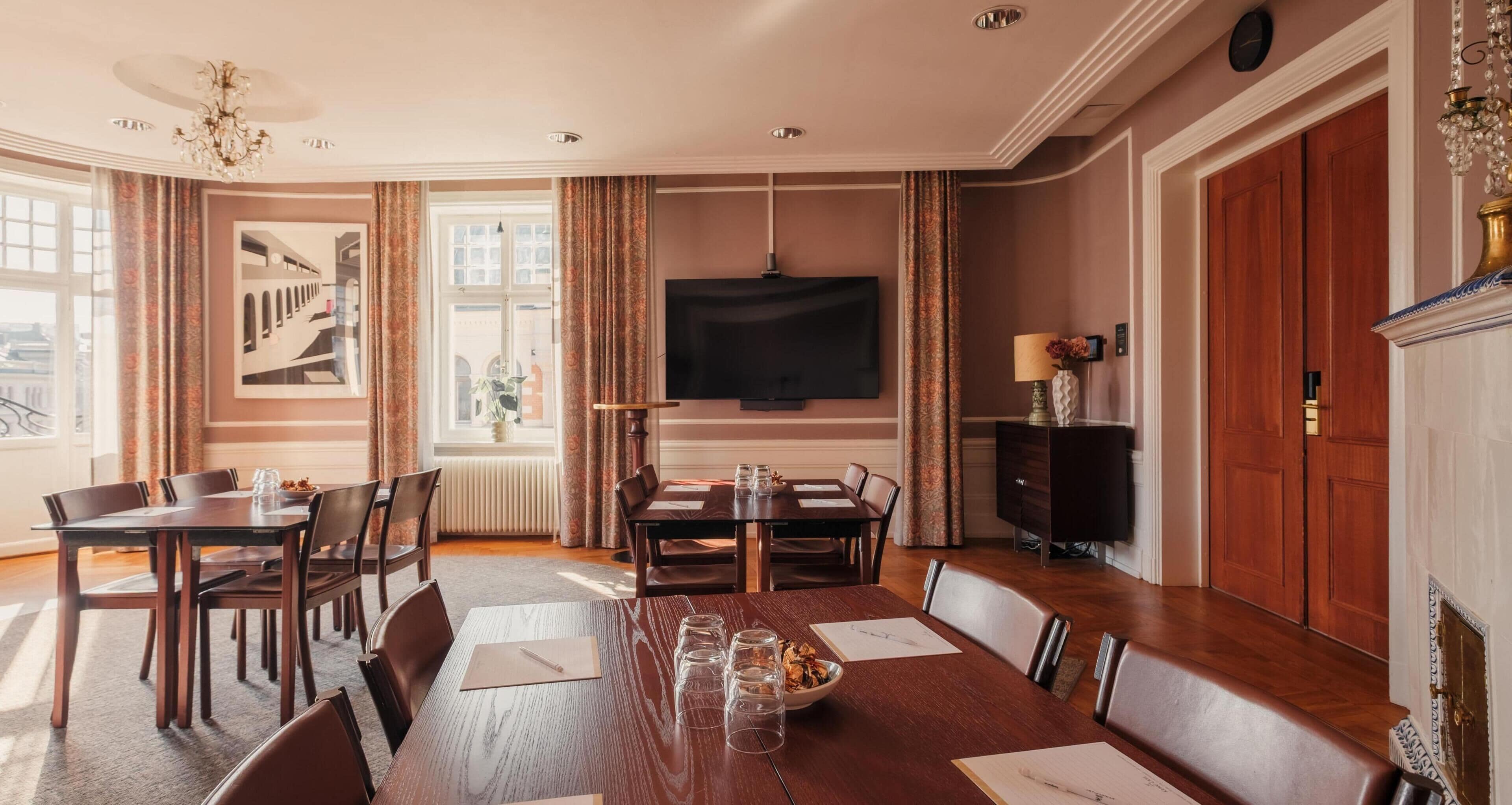 A dining room with a large table, chairs, a chandelier, a television, and a view of a building through a window.