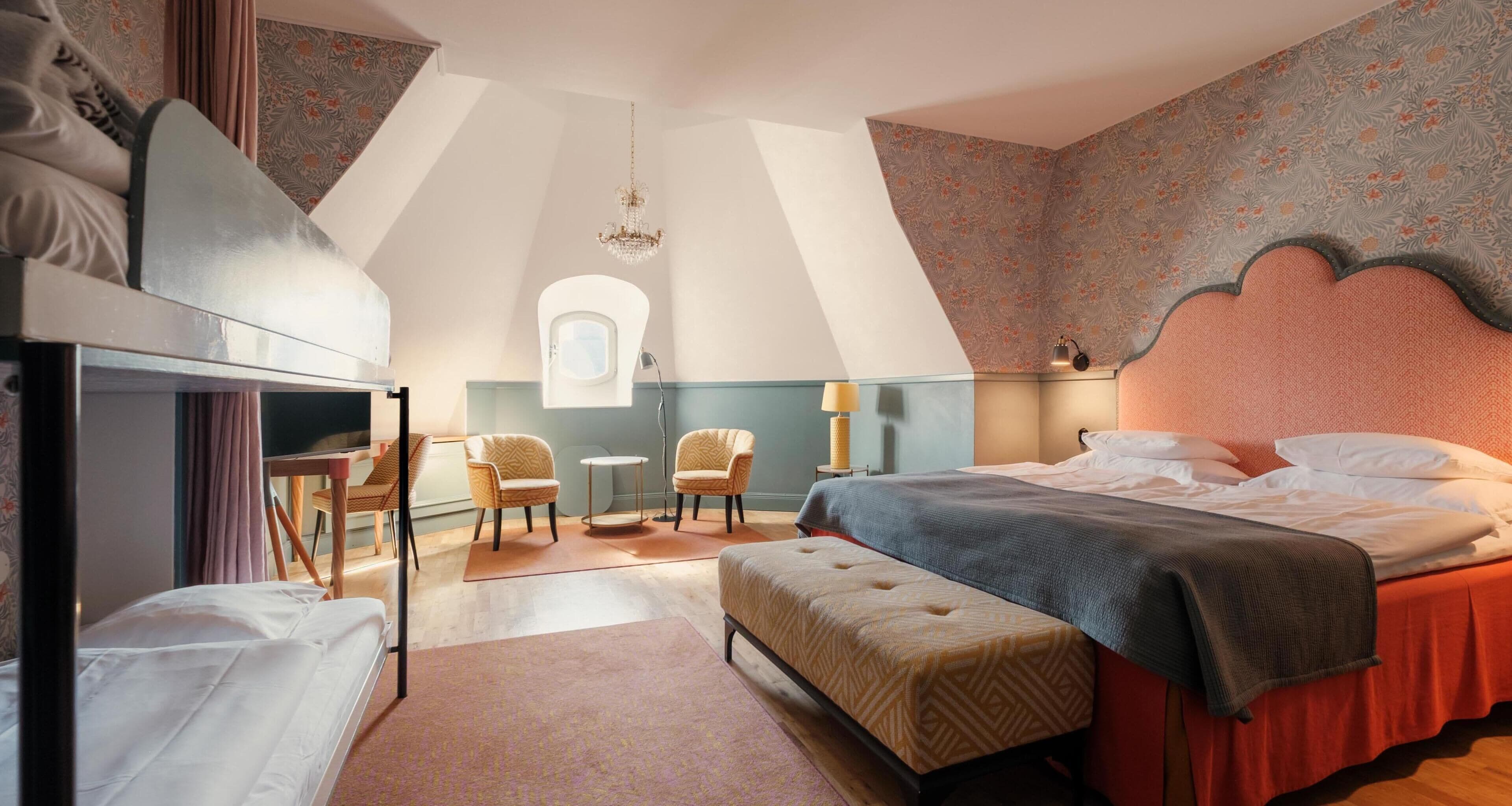 A bedroom with a bed, two chairs, a desk, a small table, a window, and a chandelier.